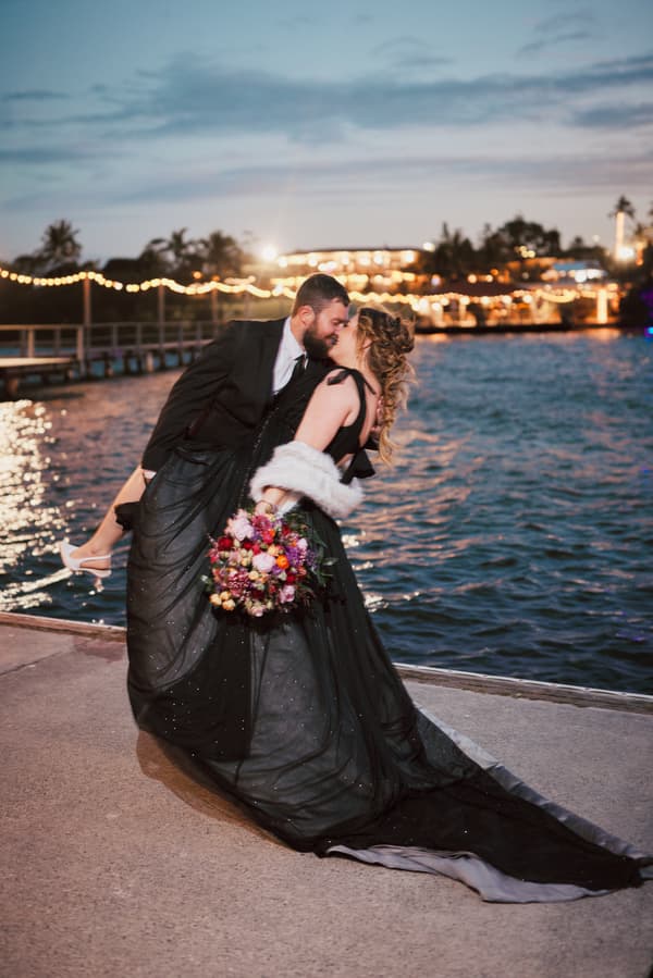 The bride Jayme in a black gown holding a colorful bouquet is dipped backward by the groom Christopher in a black suit near the water at Sandstone Point Hotel during twilight.