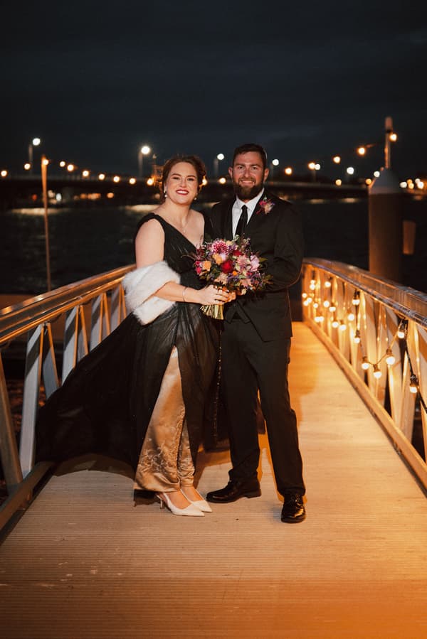 The bride Jayme and groom Christopher stand together on a lit pier at night, Jayme holding a colorful bouquet and wearing a black dress with a white shawl, Christopher in a black suit with a boutonniere.
