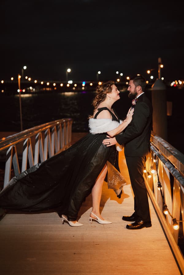 The bride Jayme in a black gown with a white shawl and the groom Christopher in a black suit stand facing each other on a lit pier at night with water and distant lights in the background.