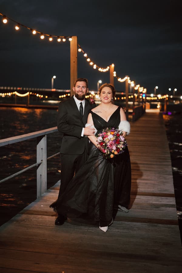 Bride Jayme in a black gown holding a bouquet and groom Christopher in a black suit stand together on a wooden pier decorated with string lights at night.