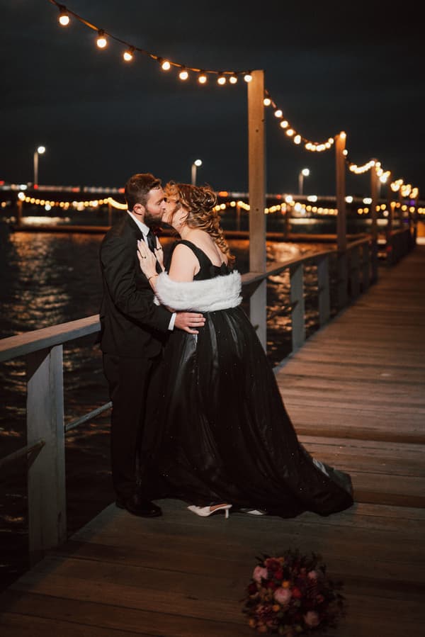 Jayme and Christopher share a kiss on a wooden pier at night, decorated with string lights, with water visible in the background and a bouquet on the ground nearby.