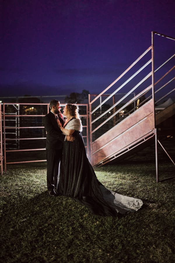 The bride Jayme and groom Christopher stand facing each other on grass at night near metal railings and a ramp structure at Sandstone Point Hotel.