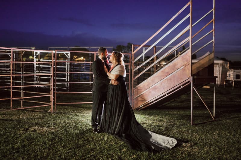 The bride Jayme in a dark gown with a white fur stole and the groom Christopher in a black suit stand closely together on grass at dusk near metal fencing and a staircase structure.