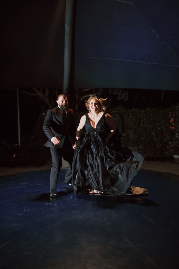 The bride Jayme and groom Christopher hold hands and walk together on a dark floor at night, with Jayme wearing a flowing black dress and Christopher in a black suit at Sandstone Point Hotel — The Cellar.