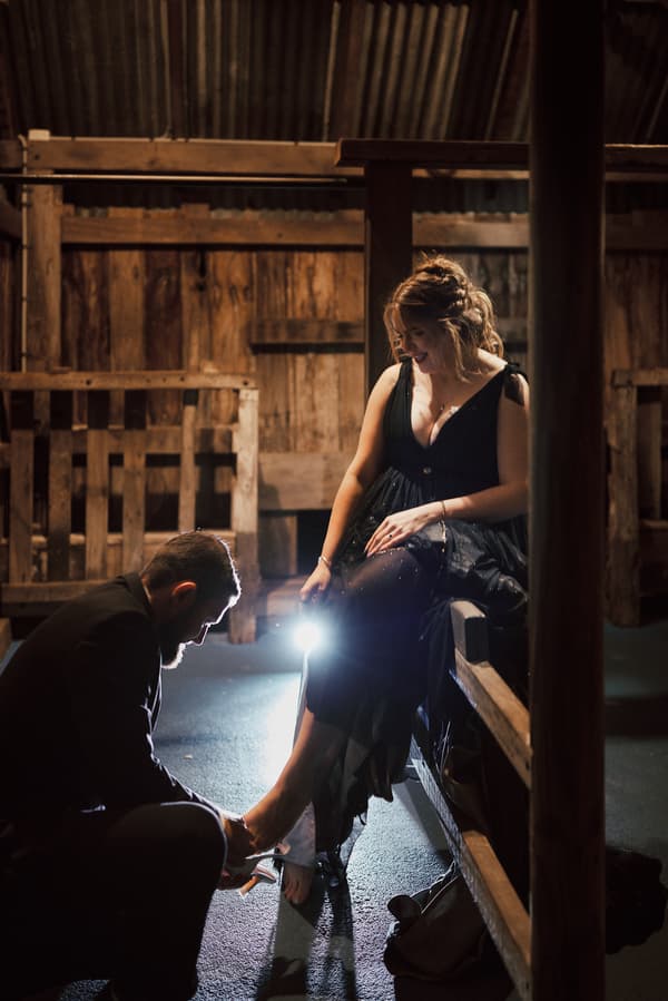 The groom kneels to help the bride put on her shoe inside a rustic wooden structure at Sandstone Point Hotel — The Pavilion.
