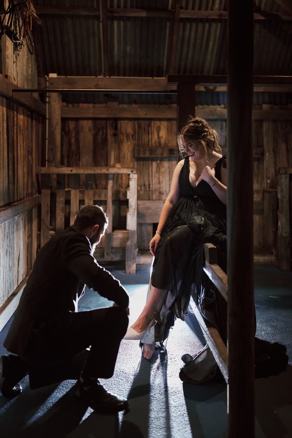 The bride sits on a wooden bench inside a rustic wooden structure at Sandstone Point Hotel, The Pavilion, while the groom kneels in front of her, helping with her shoe.