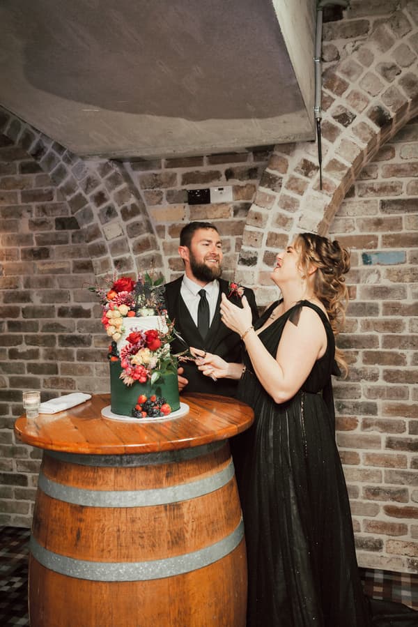 Bride Jayme and groom Christopher stand together behind a decorated wedding cake on a wooden barrel table at Sandstone Point Hotel — The Cellar reception stage.