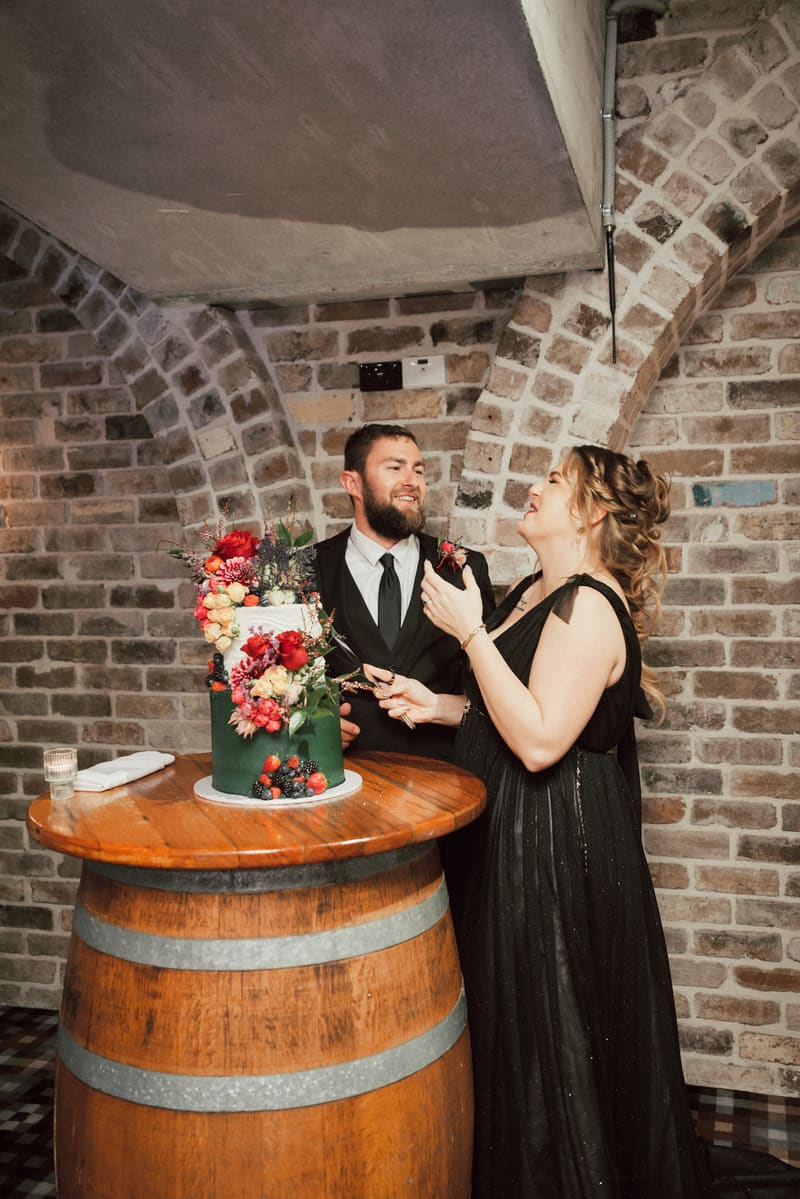 Bride Jayme and groom Christopher stand together behind a decorated wedding cake on a wooden barrel table. Easter long weekend wedding Brisbane by She Said Yes.