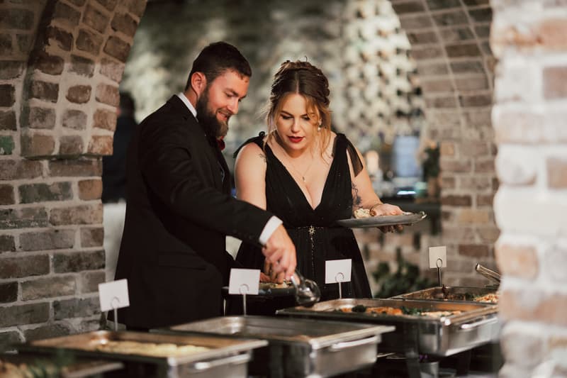 Bride Jayme and groom Christopher serve themselves food from a buffet at the reception stage of Sandstone Point Hotel — The Cellar.