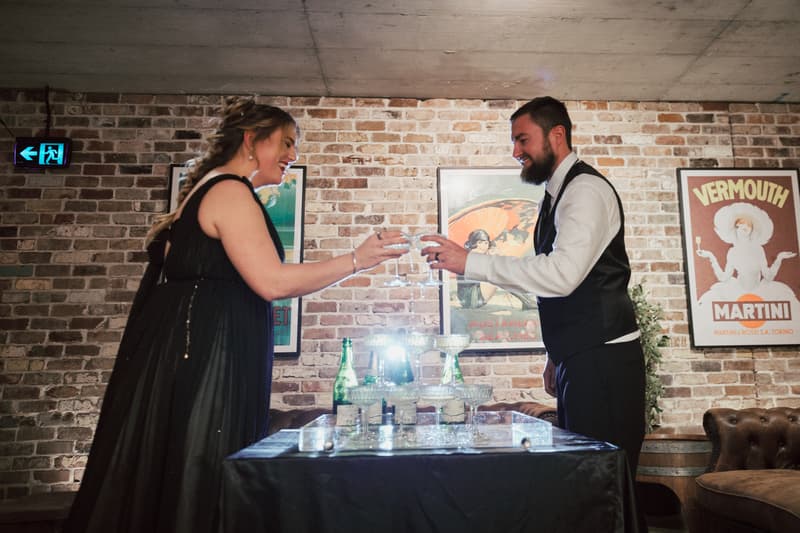 The bride Jayme and the groom Christopher toast with champagne glasses at a table with a champagne tower inside Sandstone Point Hotel — The Cellar.