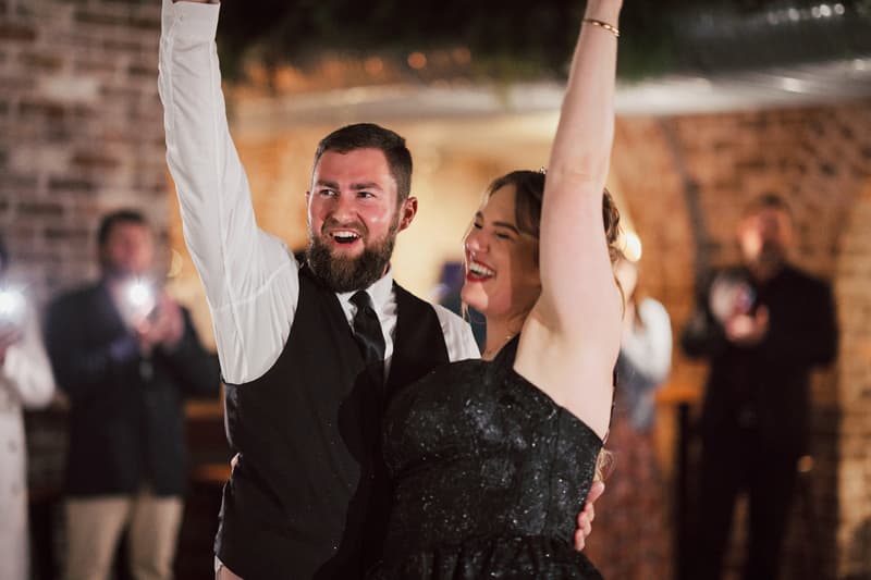 Christopher and Jayme raise their arms in celebration at the reception stage of Sandstone Point Hotel — The Cellar, with guests in the background taking photos.
