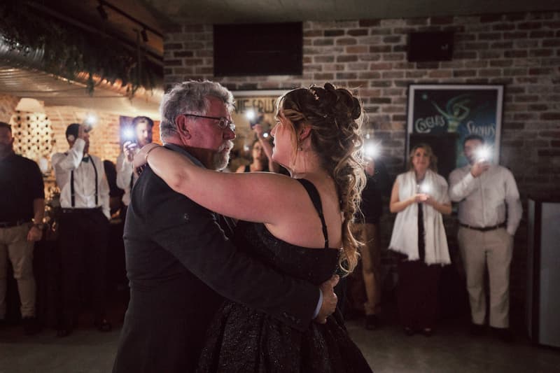 The bride Jayme dances with an older man, likely her father, at the Sandstone Point Hotel — The Cellar during the reception. Guests in the background take photos with their phones.