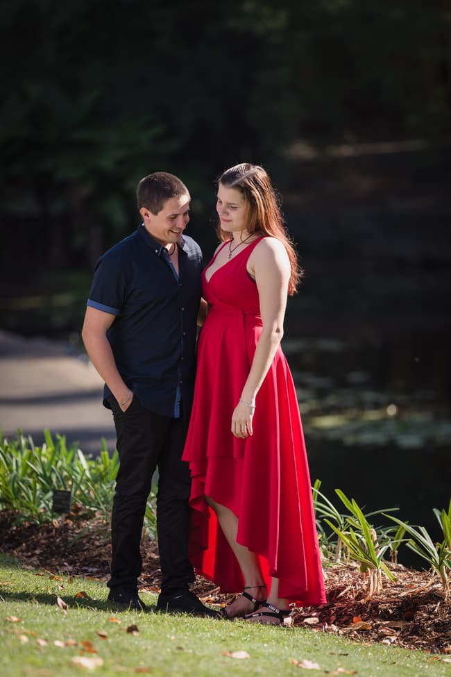 The couple stands closely on a grassy area, with the woman in a red dress and the man in a dark shirt, smiling at each other in the Brisbane Botanic Gardens.