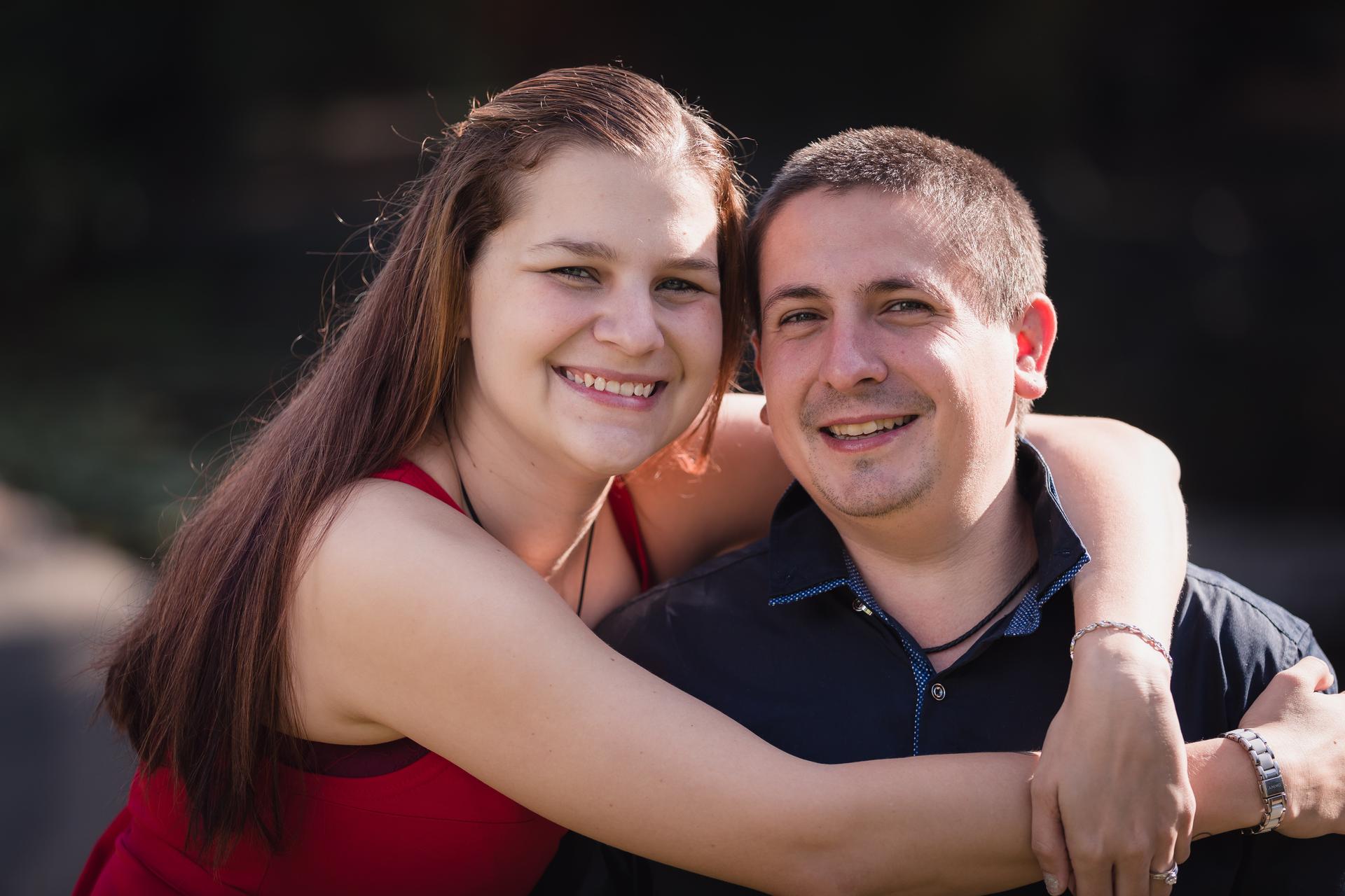 The couple poses closely, with the woman embracing the man from behind, both smiling warmly at the camera.