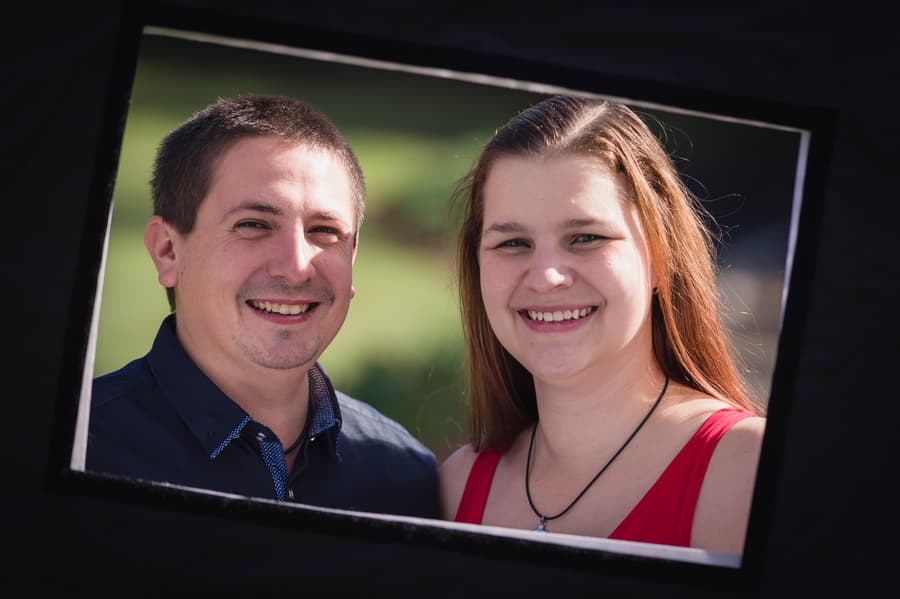 The couple smiles brightly, framed by a black photo prop, with greenery in the background.