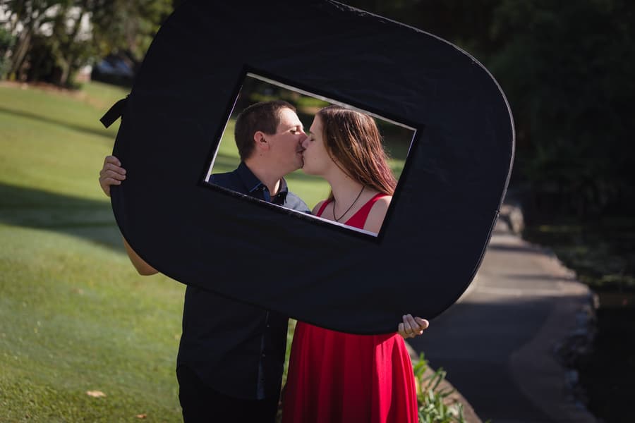 The couple kisses while holding a black frame, standing on a path with lush greenery around them.