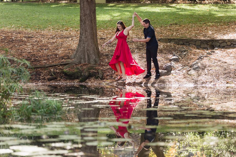 The couple dances playfully near a pond, with their reflection visible in the water, surrounded by trees.