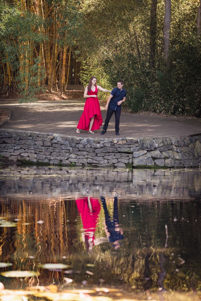 The couple stands on a stone path by a pond, with their reflection visible in the water, surrounded by bamboo.