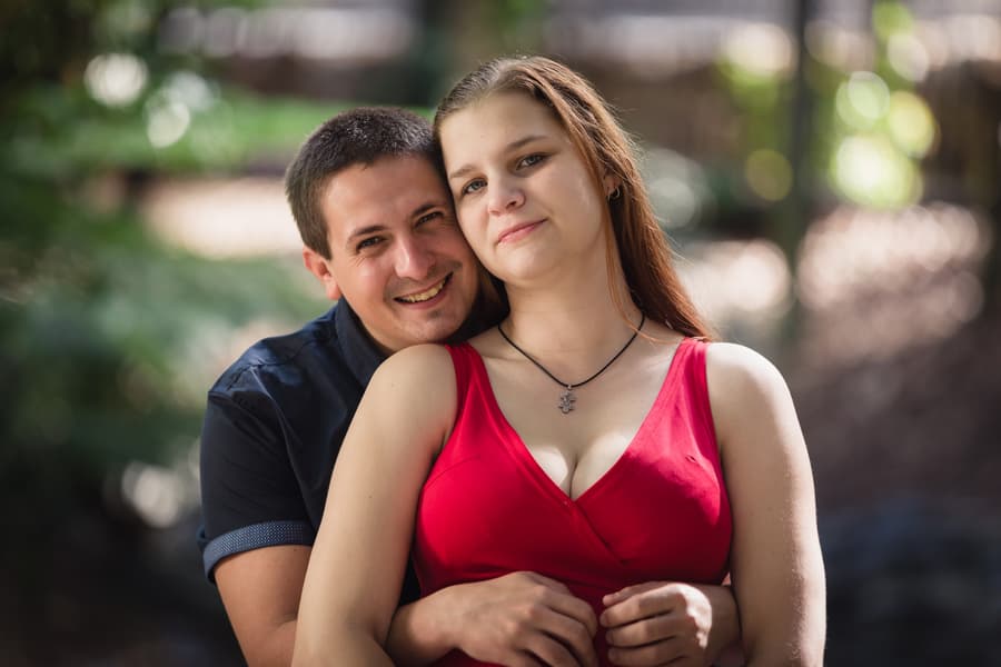 The man embraces the woman from behind, both smiling softly, with a blurred natural background.