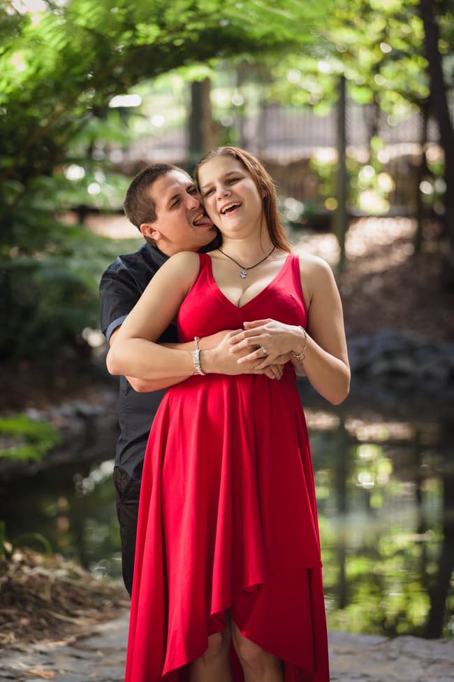 The couple laughs together, with the man playfully embracing the woman from behind, surrounded by greenery.