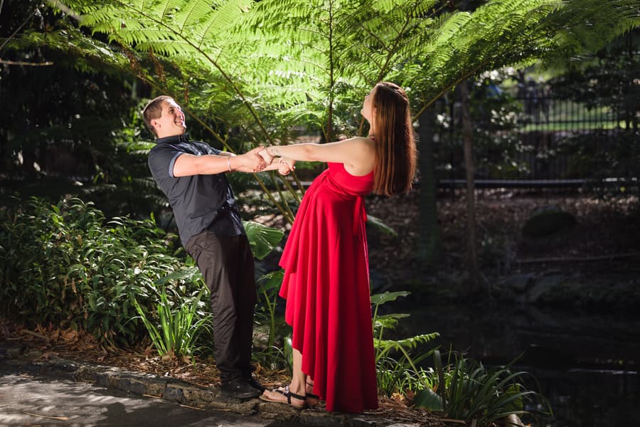 The couple playfully leans back, holding hands, surrounded by lush ferns and greenery.