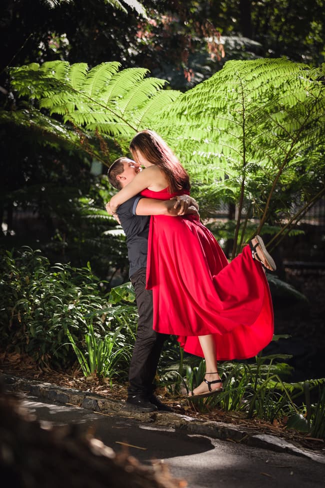 The man lifts the woman in a joyful embrace, surrounded by vibrant greenery and dappled sunlight.