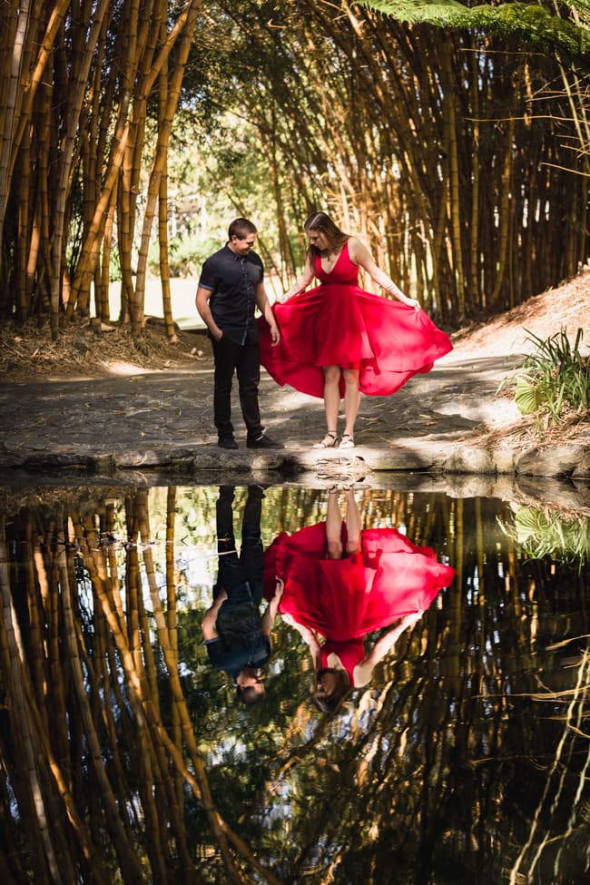 The couple stands by a reflective pond surrounded by tall bamboo at the Japanese Garden, Brisbane Botanic Gardens Mt Coot-tha. The woman in a red dress looks down at her reflection.