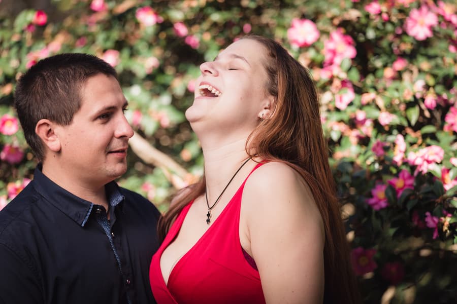 The couple stands in front of vibrant pink flowers. The woman in a red dress is laughing joyfully while the man looks at her with a smile.