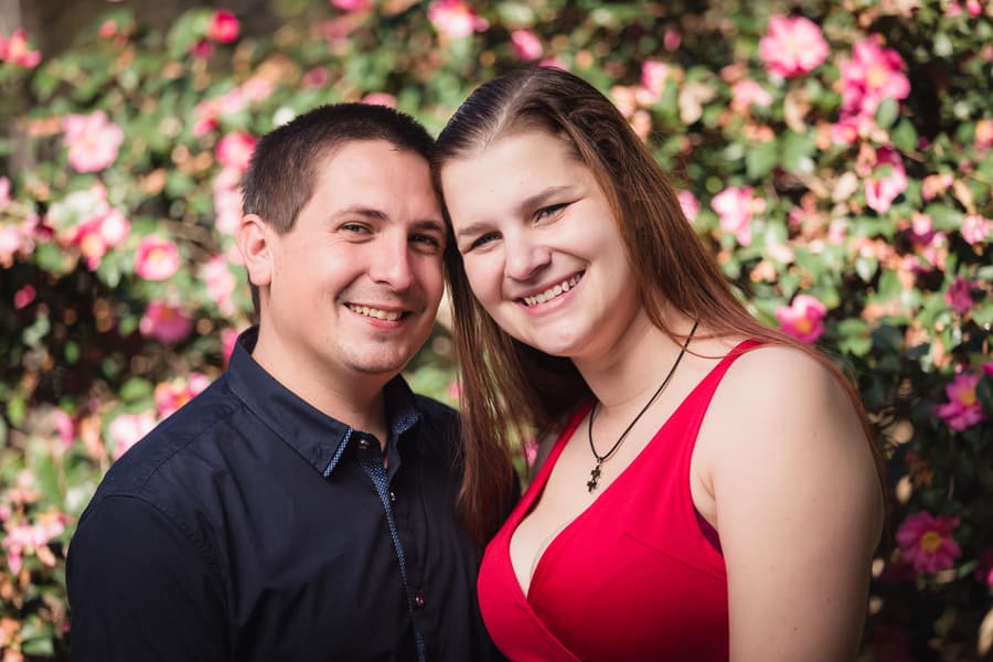 The couple stands close together, smiling warmly at the camera, with a backdrop of pink flowers.
