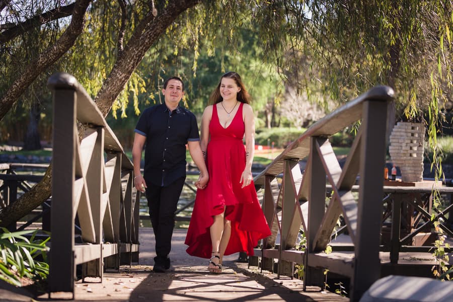 The couple walks hand in hand across a wooden bridge, surrounded by lush greenery at the Japanese Garden.