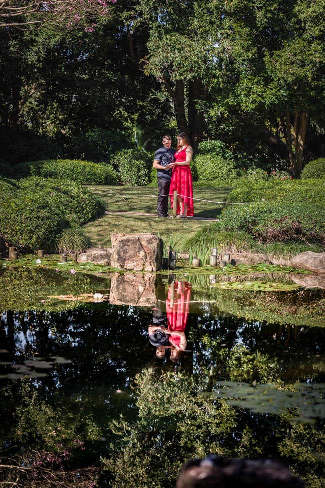 The couple stands on a grassy area with their reflection visible in a pond, surrounded by lush greenery.