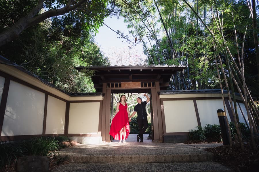 The couple walks through a traditional Japanese gate, holding hands and smiling, surrounded by greenery.