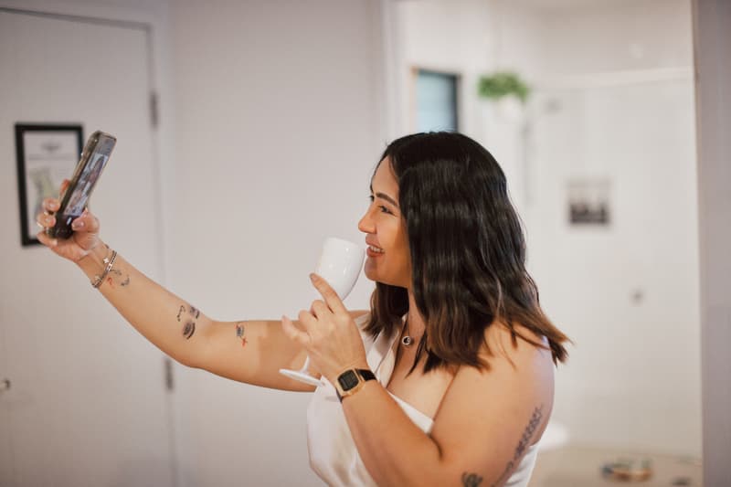 The bride takes a selfie while holding a white cup inside a room.