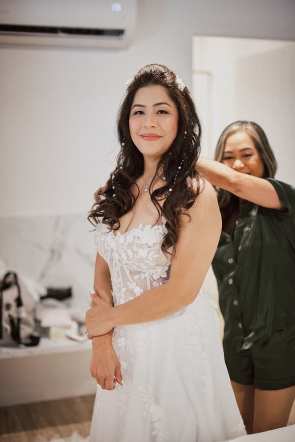 The bride Jheng stands wearing a white floral lace wedding gown while a woman in dark clothing adjusts her dress in a preparation room.