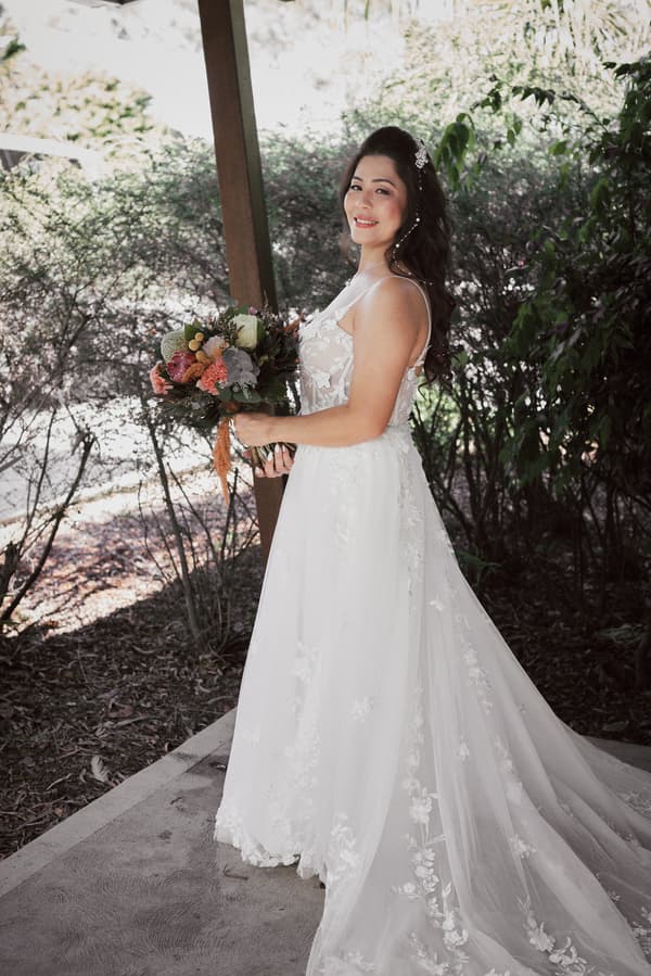 The bride Jheng stands outdoors near foliage holding a bouquet of flowers, wearing a white lace wedding gown with a long train and hair accessories.