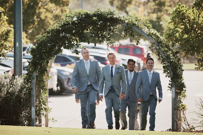 The groom Dane and three groomsmen walk under a floral arch at Sandstone Point Hotel — The Pavilion, outdoors near a parking area.