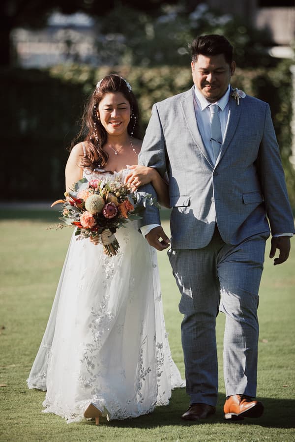 The bride Jheng in a white floral lace gown holding a bouquet walks arm-in-arm with the groom Dane in a light grey suit and tie on the grass at Sandstone Point Hotel — The Pavilion.