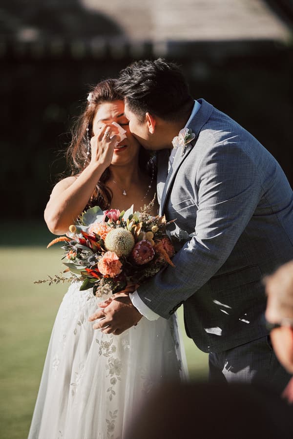The bride Jheng wipes her tears while the groom Dane leans in to kiss her cheek during their wedding ceremony at Sandstone Point Hotel — The Pavilion. Jheng holds a bouquet of mixed flowers.