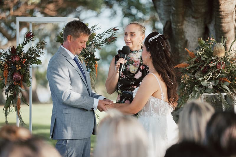 Bride Jheng and groom Dane hold hands during their wedding ceremony at Sandstone Point Hotel — The Pavilion, with the celebrant speaking into a microphone beside them.