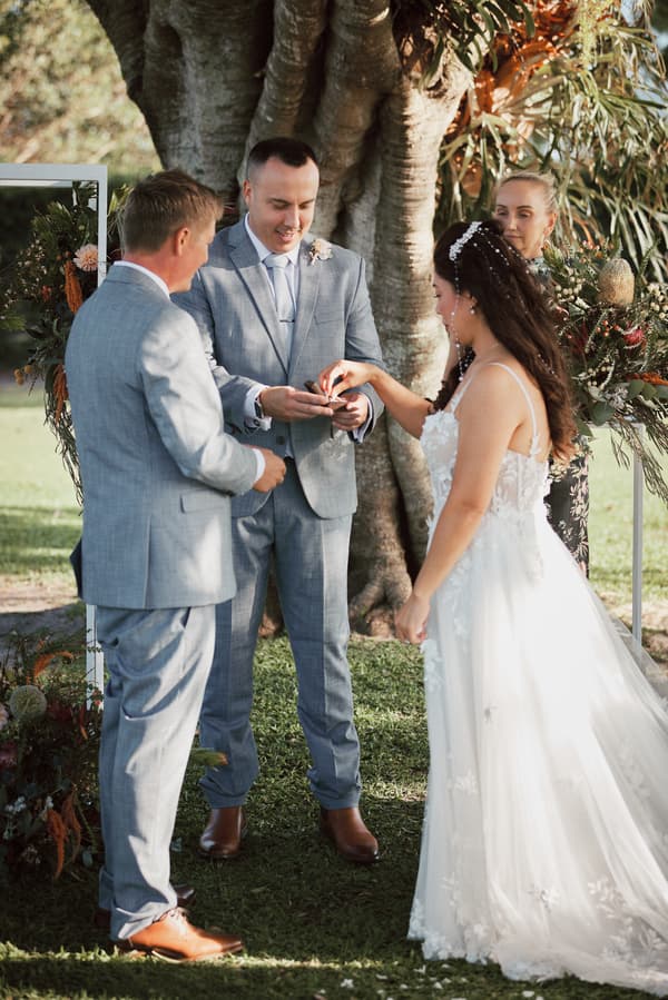 Jheng the bride places a ring on Dane the groom's finger during the wedding ceremony at Sandstone Point Hotel — The Pavilion, with the officiant and a groomsman standing nearby.