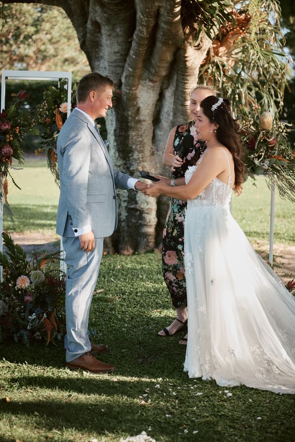 Bride Jheng and groom Dane hold hands during their wedding ceremony at Sandstone Point Hotel — The Pavilion, with the officiant standing behind them holding a microphone.