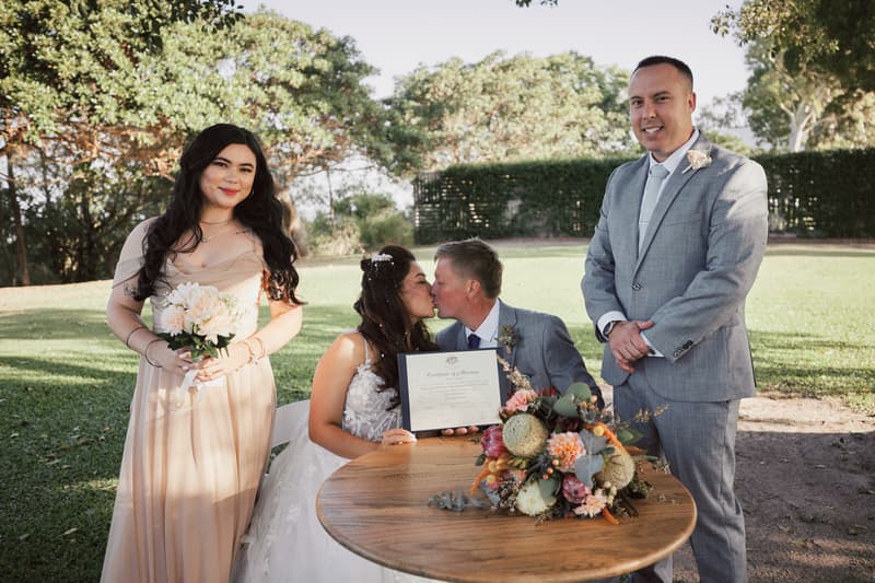 Bride Jheng and groom Dane kiss while holding their marriage certificate at a wooden table with a floral bouquet, accompanied by a bridesmaid holding a bouquet, at Sandstone Point Hotel — The Pavilion outdoors.