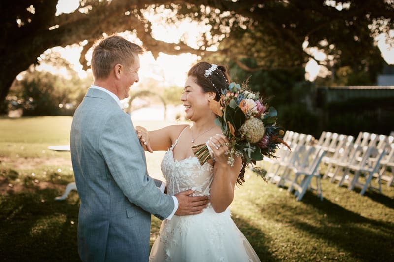 Bride Jheng and groom Dane smile at each other while embracing outdoors, with rows of white chairs visible in the background under a large tree. Engagement ceremony by She Said Yes.