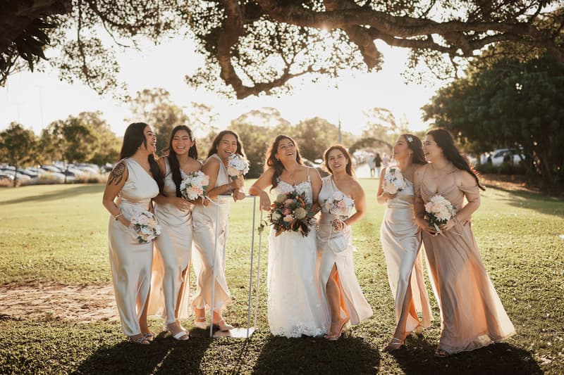 The bride Jheng stands with six bridesmaids holding bouquets on a grassy area under a large tree at Sandstone Point Hotel during the couple portraits stage.