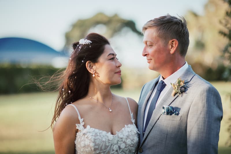 The bride Jheng and groom Dane stand facing each other outdoors at Sandstone Point Hotel, with Jheng wearing a white floral lace wedding dress and hair accessories, and Dane in a light grey suit with a boutonniere and blue tie.