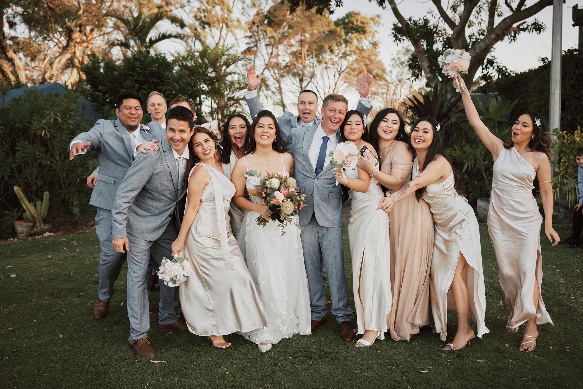 Bride Jheng and groom Dane pose with their wedding party outdoors at Sandstone Point Hotel, surrounded by bridesmaids in light-colored dresses and groomsmen in grey suits.