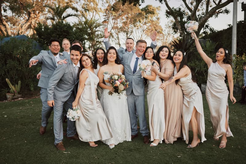 Bride Jheng and groom Dane pose with their wedding party outdoors at Sandstone Point Hotel, surrounded by bridesmaids in light-colored dresses and groomsmen in grey suits.