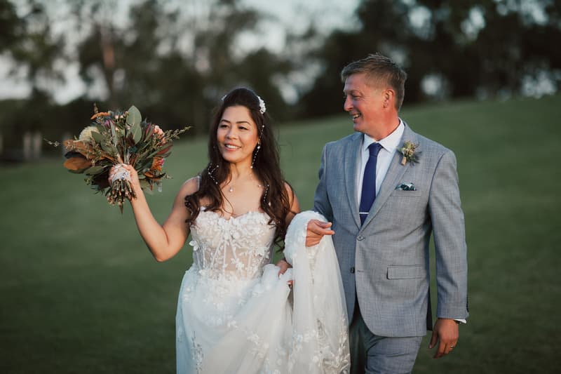 The bride Jheng holds her bouquet and lifts her wedding dress while walking beside the groom Dane on a grassy area at Sandstone Point Hotel.