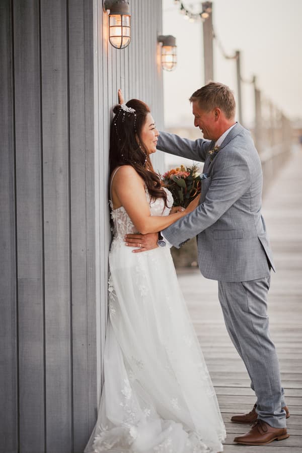 The bride Jheng in a white lace wedding gown and the groom Dane in a light grey suit pose together on a wooden boardwalk at Sandstone Point Hotel. Jheng is leaning against a grey wooden wall with lit wall lamps, holding a bouquet, while Dane faces her with one arm resting on the wall.