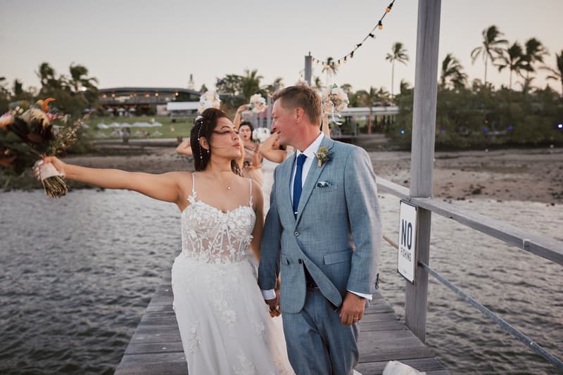 The bride Jheng and groom Dane walk hand in hand on a wooden pier at Sandstone Point Hotel. Jheng holds a bouquet out to the side while looking at Dane, who is dressed in a light grey suit with a blue tie. Bridesmaids are visible in the background on the pier.